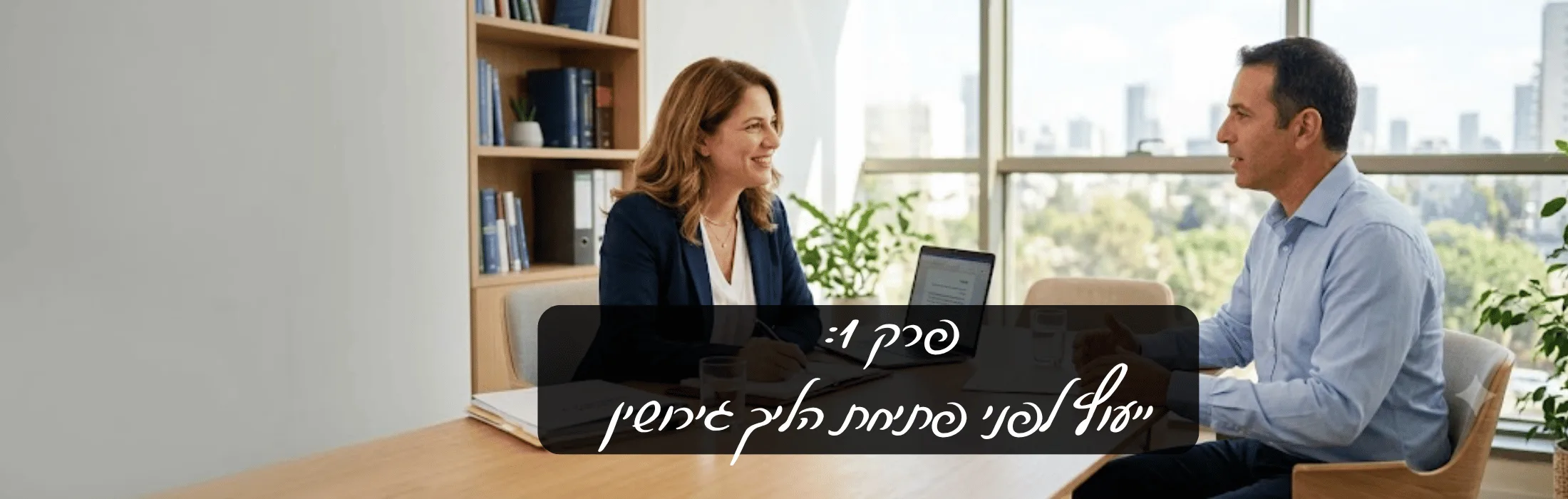 Two coworkers discuss at a wooden table in a bright office, laptop open, with a dark overlay bearing Hebrew text across the center.