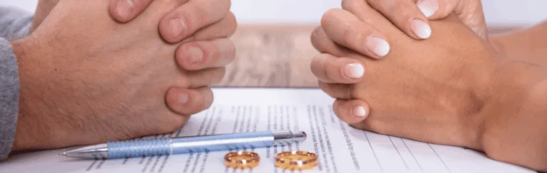 Two people clasping hands over a signed document with a blue pen and wedding rings nearby.
