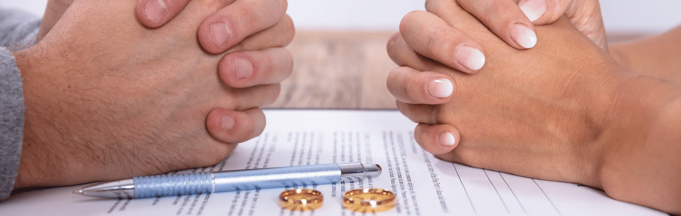 Two people clasping hands over a signed document with a blue pen and wedding rings nearby.